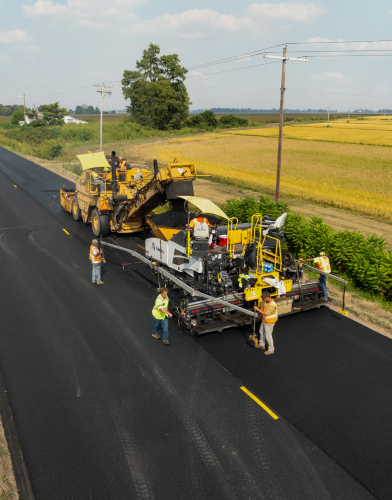 Construction vehicles working on a rural road surrounded by open fields.