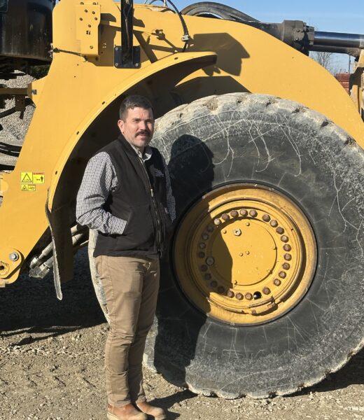 A construction worker wearing a vest and leaning against a large industrial tire, smiling.